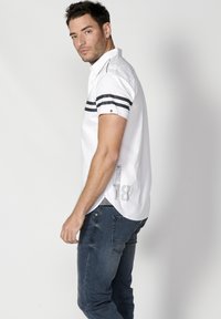 Camisa blanca de manga corta con rayas negras en las mangas; presenta una etiqueta en el costado y un dobladillo curvado. Combinada con unos jeans de mezclilla oscura.