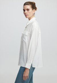 Camisa blanca de manga larga en tejido ligero con cuello clásico, bolsillo frontal y puños abotonados, combinada con jeans azules.