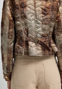 Quiltet jakke i brune og beige nuancer med et geometrisk mønster, kendetegnet ved en bæltestrop i taljen og ribbet struktur, parret med beige bukser.