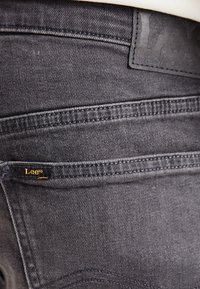 Jeans de mezclilla gris oscuro con un parche de cuero que presenta el logo de "Lee", costuras visibles y una textura suave.