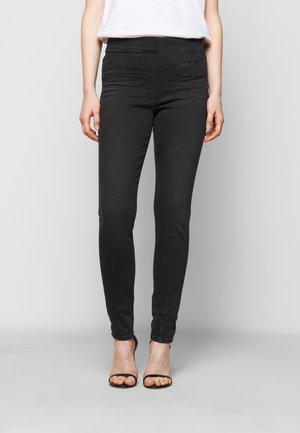 Vrouw draagt zwart nauwsluitende broek en witte top, staand met voeten op schouderbreedte uit elkaar in zwarte open-toe hakken tegen een effen achtergrond.