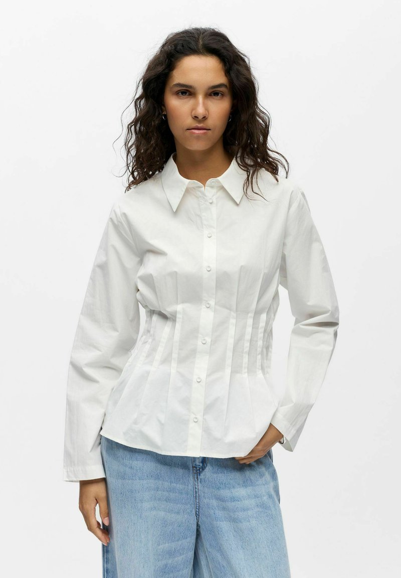 Femme aux cheveux bouclés foncés portant une chemise blanche ajustée à boutons et un jean bleu clair, debout devant un fond blanc uni.