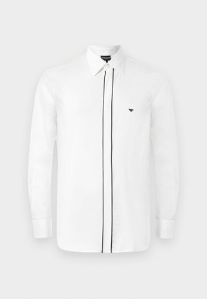 Camisa branca de manga longa com colarinho pontiagudo, apresentando um acabamento em preto ao longo da frente e um pequeno logótipo preto no peito. Textura suave.