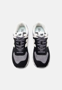 Pár čiernych, sivých a bielych tenisiek New Balance 574 so zviazanými šnúrkami, pozorované zhora na bielom pozadí.