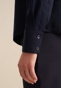 Marineblaue Langarmbluse mit Knopfmanschette, die eine glatte Textur und einen taillierten Schnitt aufweist. Sie wird vor einem neutralen Hintergrund präsentiert.