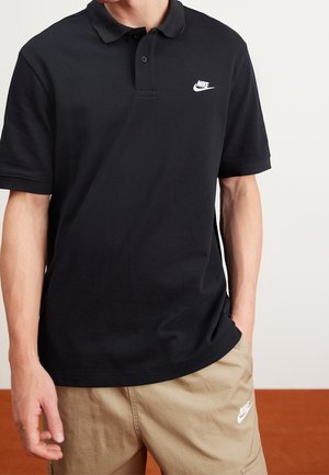 Personne portant un polo noir Nike et un short beige Nike, debout avec une main dans la poche devant un fond blanc et marron uni.