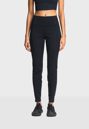 Femme portant des leggings noirs taille haute avec poches latérales et chaussures de sport noires, debout devant un fond clair uni.