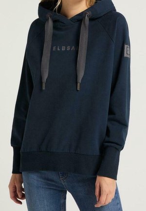 Vrouw draagt een marineblauwe hoodie met grote grijze trekkoorden, de tekst "ELBSAND" op de borst en een patch-logo op de linker mouw, gecombineerd met een blauwe spijkerbroek.