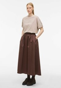 Beżowa bawełniana koszulka z wytłoczonym logo "ROUGE EDIT" w połączeniu z długą, ciemnobrązową spódnicą w stylu boho, z bocznymi kieszeniami i warstwową fakturą.