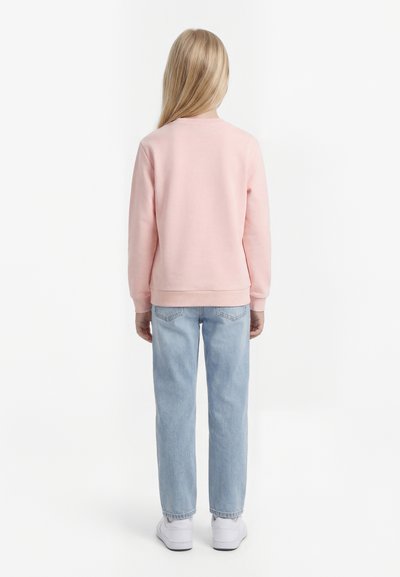 Enfant aux longs cheveux blonds portant un sweat-shirt rose pastel, un jean bleu clair et des baskets blanches, debout de dos sur un fond blanc.