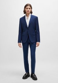 Marineblauer Anzug mit tailliertem Schnitt, einreihige Jacke, Reverskragen und schmalen Hosen. Kombiniert mit einem weißen Hemd und schwarzen Schuhen.