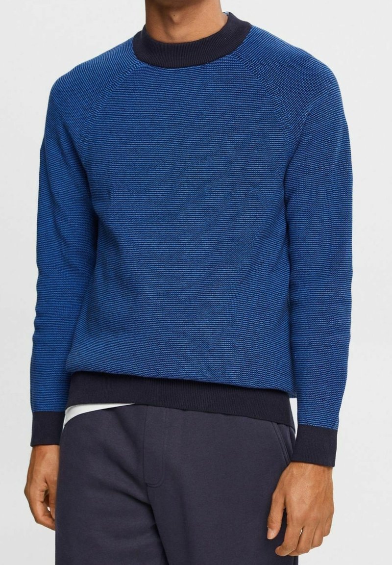 Blå stickad tröja med texturerade ränder, ribbad marinblå krage och manschetter. Har en rundad halsringning och en avslappnad passform, tillsammans med mörka byxor.