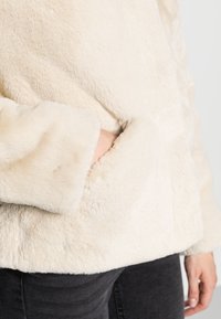Cream faux fur jacket with a soft, textured surface and hidden side pocket. Features wide sleeves and a relaxed fit. Dark jeans visible below.