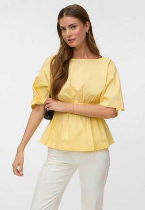 Femme aux longs cheveux bruns porte un haut jaune ample avec des manches bouffantes et un pantalon blanc, tenant un petit sac noir à sa taille.