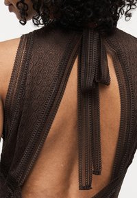 Γυναίκα με σγουρά μαλλιά που φοράει αμάνικο σκούρο καφέ δαντελένιο φόρεμα με ανοιχτή πλάτη και δέσιμο στο λαιμό με δαντελένιους ιμάντες.