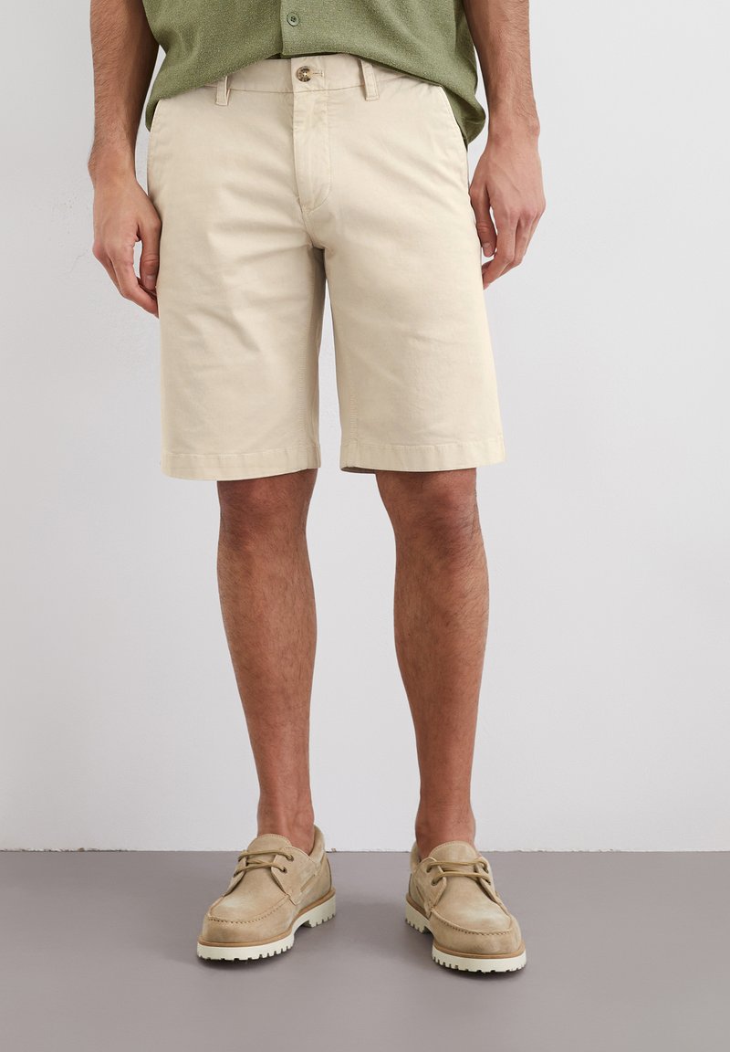 Beige cotton shorts with a straight cut, featuring a button closure and belt loops. Worn with beige footwear and a green shirt.