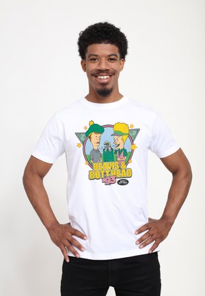Lachende man met krullend haar die een wit Beavis & Butt-Head '93 grafisch T-shirt draagt, staat met de handen in de zij tegen een effen achtergrond.