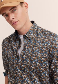 Braunes Blumenhemd mit blauen und weißen Akzenten, mit Knopfleiste, aus weichem Stoff und Kragen, über einem hellgrauen T-Shirt getragen.