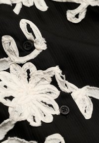 Tissu noir à texture côtelée décoré d'appliques florales en tissu blanc et de petits boutons noirs avec du texte autour des bords.