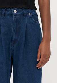 Denim trousers in dark blue with a relaxed fit, featuring pleats, a front button fastening, and side pockets. Textured fabric visible.