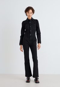 Črn denim kombinezon z oprijetim krojem, dolgimi rokavi, gumbom na sprednji strani, žepi na prsih in razširjenimi hlačami; obut z črnimi čevlji.