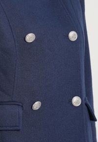 Ναυτικό διπλής σειράς παλτό από υφασμάτινη υφή, με τέσσερα ασημένια κουμπιά και δομημένο σχέδιο με πλαϊνές τσέπες.