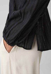 Blusa nera in tessuto testurizzato, con motivi a zigzag e polsini con bottoni. Indossata con pantaloni di colore crema, che mostrano dettagli delle tasche laterali.