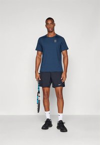 Marineblå atletisk t-skjorte med korte ermer og en liten logo, kombinert med svarte shorts. Personen holder en tennisracket og har på seg svarte sko.