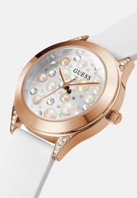 Roosa kuldne kell valgel rihmal, kaunistatud pärlite ja kristallidega kell face'il ja lukkudel, näidates Guess brändi dial'il.