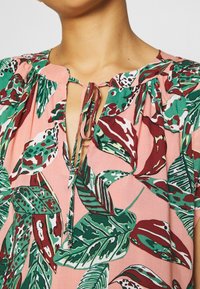 Blusa floreale con sfondo rosa e motivi di foglie verdi e bordeaux, scollatura a chiave e chiusura con fiocco. Tessuto leggero e fluido.