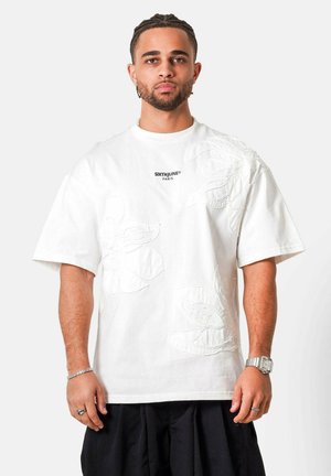 Man met gevlochten haar, draagt een wit oversized T-shirt met bloemrijke borduurwerk en de tekst "SIXTHJUNE PARIS", zwarte broek en zilveren accessoires.