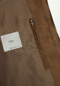 Intérieur d'une veste marron montrant une étiquette blanche MNG Man et une petite poche zippée du côté droit.