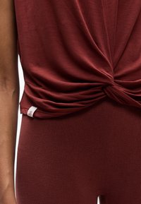 Gros plan sur une personne portant un haut bordeaux noué et un legging assorti côtelé, montrant la texture du tissu et une petite étiquette blanche.
