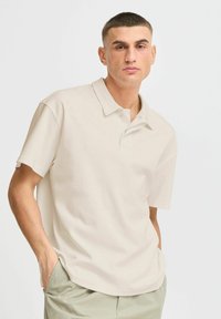 Polo shirt beige clair avec une coupe décontractée, des manches courtes, un col et une finition texturée subtile. Présente deux boutons sur la patte de boutonnage.