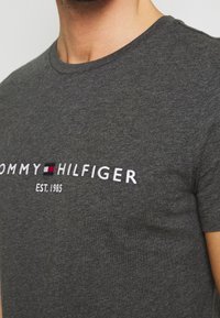 Primer plano de una persona que lleva una camiseta gris oscura de Tommy Hilfiger con un logo bordado en blanco y el texto "EST. 1985" en el pecho.