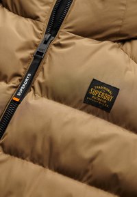 Giacca a vento marrone con zip nera, sezioni imbottite texturizzate e un patch con logo in giallo e bordatura nera.