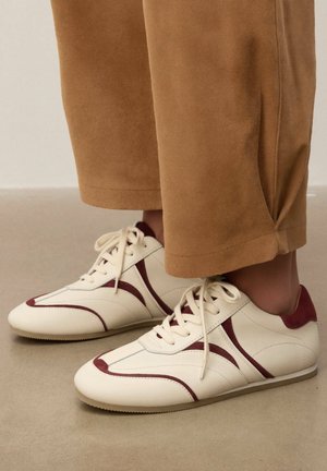 Witte en bordeauxrode veter sneakers gedragen met losse tan broek op een neutrale vloer, met zijkant- en voorkantdetails van de schoen zichtbaar.