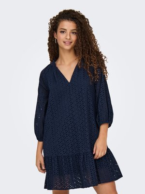 Jeune femme aux cheveux bouclés portant une robe à œillets bleu marine avec un col en V, des manches ballon trois-quarts et un ourlet à volants sur fond blanc.