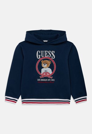 Granatowa bluza z kapturem z paskowanymi mankietami i dołem, z misiem w czerwonym oplecionym kółku oraz napisem "GUESS" powyżej, a poniżej napis Los Angeles est. 1981.