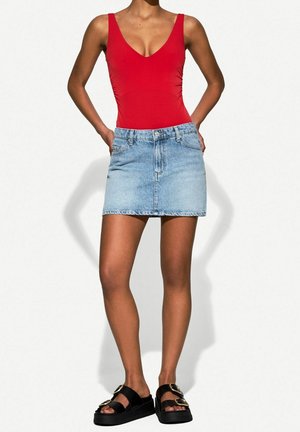 Femme portant un haut rouge sans manches, une mini-jupe en jean bleu clair et des sandales compensées noires avec des détails de boucle dorée.