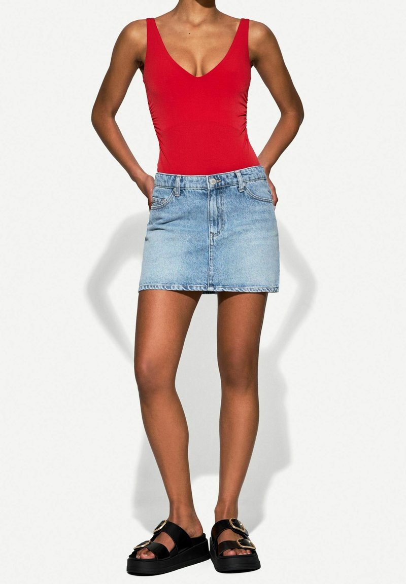 Femme portant un haut rouge sans manches, une mini-jupe en jean bleu clair et des sandales compensées noires avec des détails de boucle dorée.