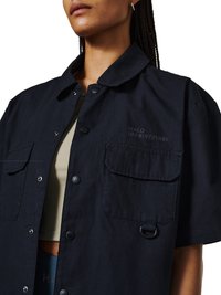 Kvinde iført en mørk marineblå skjorte med korte ærmer, to forlommer med klap og diskret tekst "HALO 196819721981" på brystet.