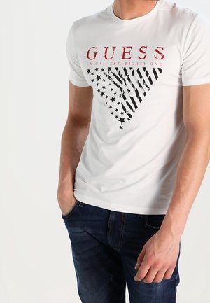 Homme portant un t-shirt blanc GUESS avec un logo triangle stylisé du drapeau américain et un jean bleu foncé, mains partiellement dans les poches.