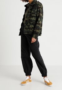 Giacca camouflage in verde scuro e nero, abbinata a pantaloni jogger neri e sneakers bianche con dettagli gialli. Quattro tasche frontali visibili.