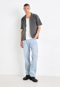 Camisa de botones de manga corta gris sobre una camiseta sin mangas blanca, combinada con jeans rectos de pierna ligera en azul claro y botas negras hasta el tobillo. Diseño simple.