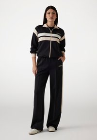 Crna atletska zip-up jakna s kremastim prugama, u paru s odgovarajućim crnim hlačama koje imaju logotip. Mekani, lagani materijal, opušten fit.