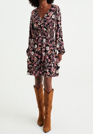 Vrouw draagt een zwarte, lange bloemenjurk met lange mouwen en een V-hals, gecombineerd met bruine suède laarzen tot aan de knie, staand tegen een effen witte achtergrond.