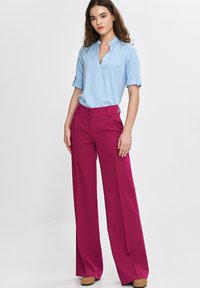 Jasnoniebieska bluzka z krótkim rękawem, z kołnierzykiem i podwiniętymi mankietami, zestawiona z fioletowymi spodniami o wysokim stanie i szerokich nogawkach oraz beżowymi butami.
