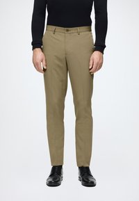 Tan broek van gladde stof, met een slim fit, knoopsluiting en frontzakken. Draagt een zwart paar schoenen en een aansluitend zwart topje.