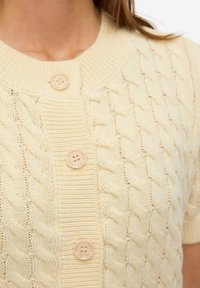 Crème gebreide cardigan met een gestructureerd kabelpatroon, ronde hals en drie grote lichtgekleurde knopen aan de voorkant. Korte mouwen.